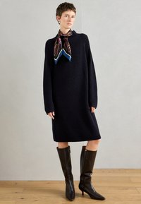 Μπλε πλεκτό φόρεμα με μακριά μανίκια, φορεμένο με μια διακοσμημένη μαντήλα. Συνδυάζεται με γυαλιστερά καφέ μποτάκια με ψηλό τακούνι μέχρι το γόνατο. Φόντο από ξύλινο πάτωμα.