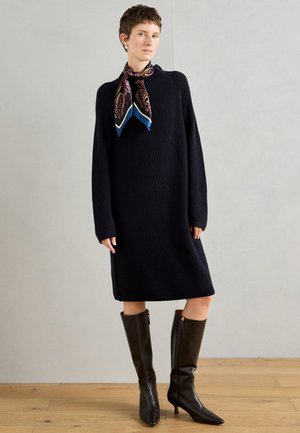 Marinblå stickad klänning med långa ärmar, bärs med en mönstrad halsduk. Sammanställd med knähöga, glansiga bruna stövlar med klack. Trägolvsbakgrund.