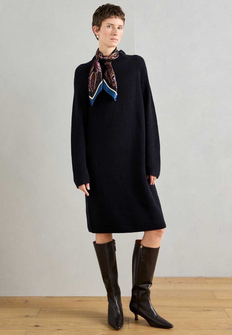 Μπλε πλεκτό φόρεμα με μακριά μανίκια, φορεμένο με μια διακοσμημένη μαντήλα. Συνδυάζεται με γυαλιστερά καφέ μποτάκια με ψηλό τακούνι μέχρι το γόνατο. Φόντο από ξύλινο πάτωμα.