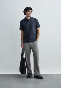 Námornícka modrá polokošeľa s jemným vzorom, sivé nohavice s širokými nohavicami a čierne tenisky. V ruke drží čiernu tašku na rameno. Čistá, moderná estetika.