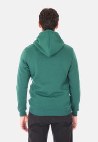 Uomo con capelli scuri e corti che indossa una felpa verde semplice e pantaloni neri, in piedi di spalle contro uno sfondo bianco.
