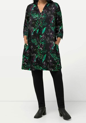 Femme portant une robe tunique noire avec un imprimé botanique vert et violet, manches trois-quarts, leggings noirs et bottines noires.