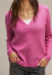 Femme portant un pull rose vif à col en V par-dessus un haut blanc orné de dentelle, les mains dans les poches d'un pantalon gris foncé.