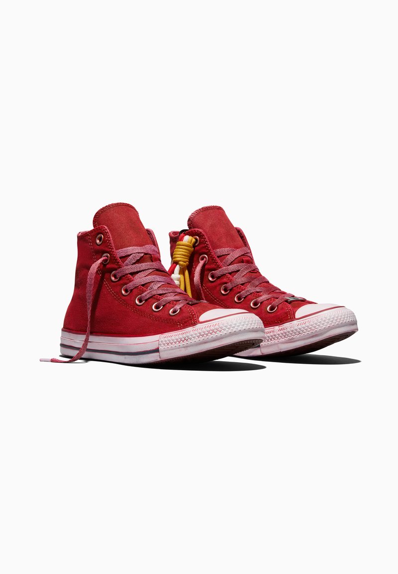 Pair of red high-top canvas sneakers with white rubber soles and pink laces, one shoe featuring a yellow decorative lace knot.
