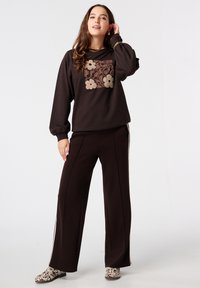 Jonge vrouw in een donkerbruin sweatshirt met bloemenborduursel en bijpassende wijde broek, met patronen enkellaarsjes op een witte achtergrond.