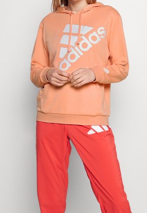 Person iført en ferskenfarvet Adidas-hoodie og røde Adidas-bukser, stående med hænderne foldet foran mod en ensfarvet baggrund.