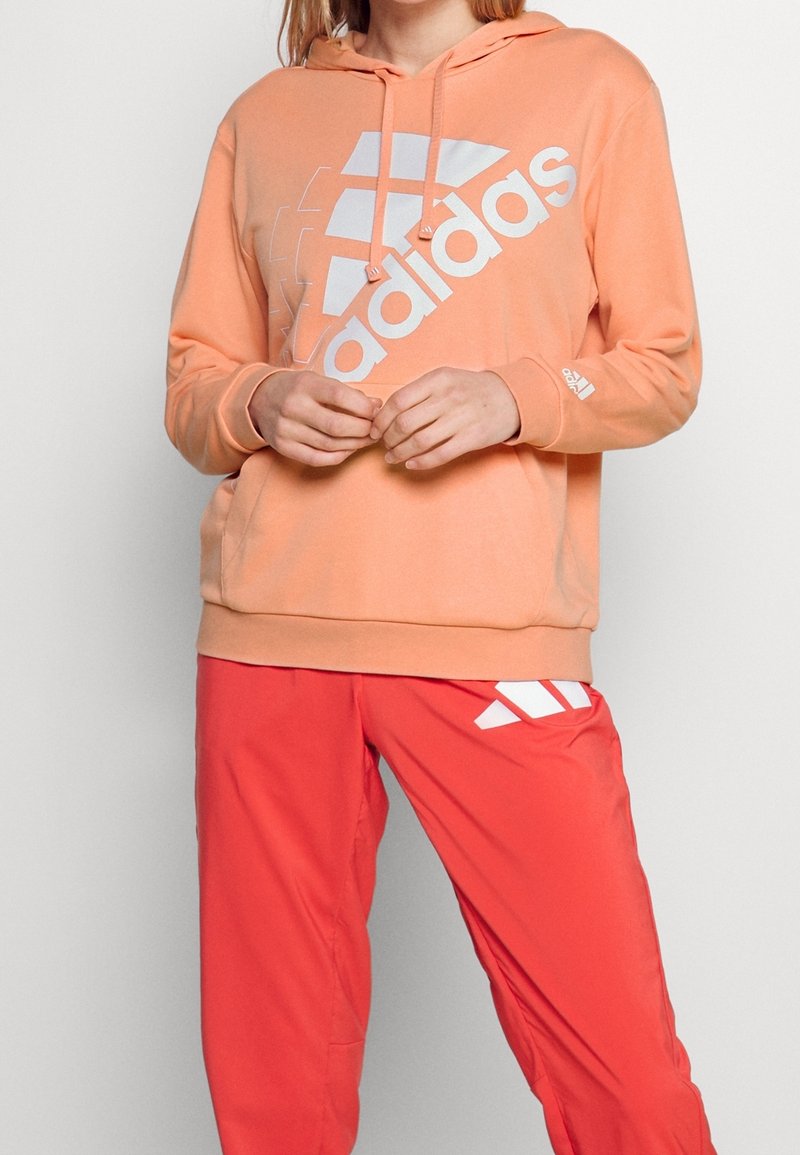 Personne portant un sweat à capuche Adidas pêche et un pantalon Adidas rouge, debout les mains jointes devant un fond uni.
