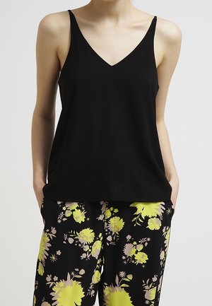 Kvinde iført en sort ærmeløs top med V-udskæring og sorte bukser med store gule og beige blomsterprint, hænder i lommerne.