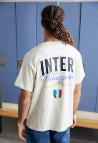 Hvit bomulls t-skjorte med "INTER" og "Nerazzurri" trykket på baksiden i fet svart og blå tekst, med dekorative elementer i gull og en logo.