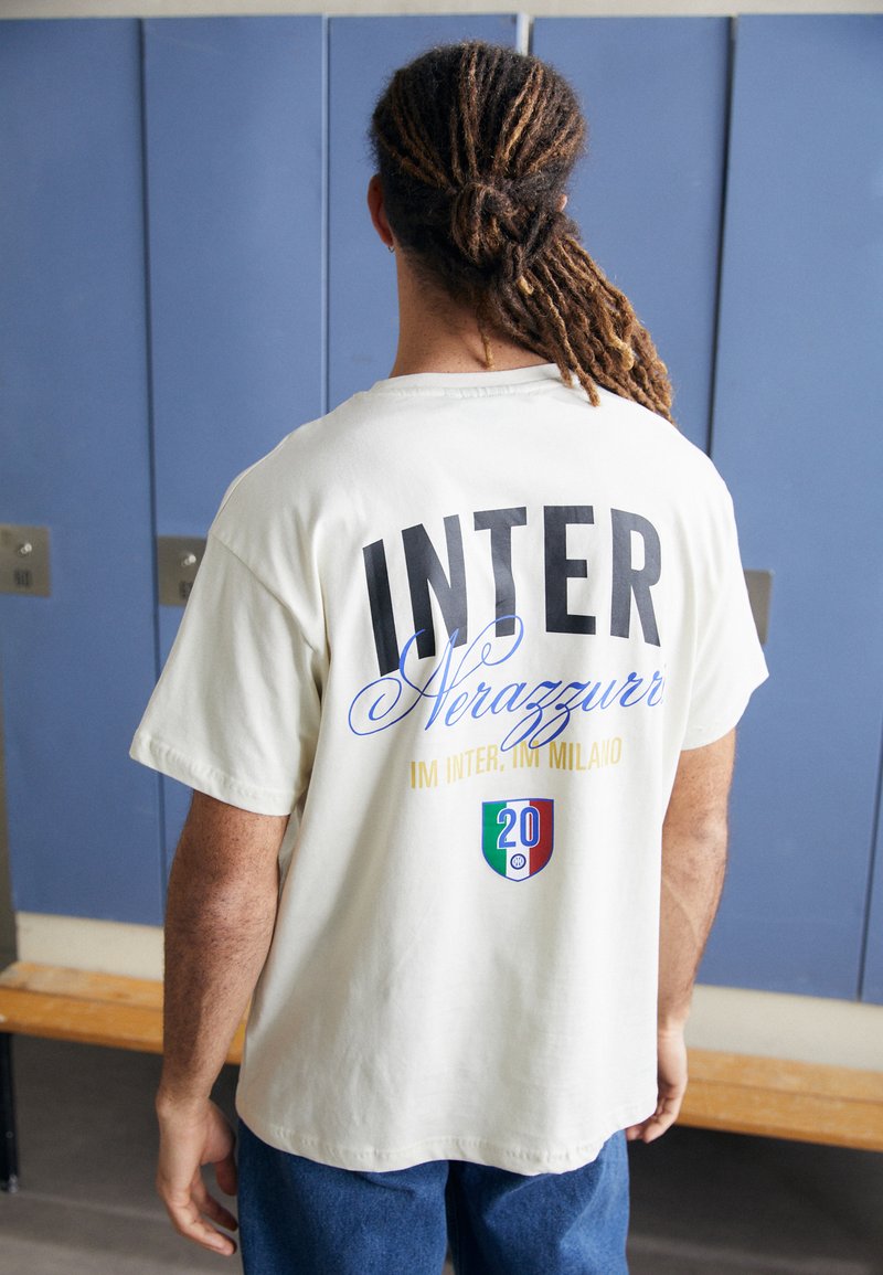 Hvit bomulls t-skjorte med "INTER" og "Nerazzurri" trykket på baksiden i fet svart og blå tekst, med dekorative elementer i gull og en logo.
