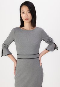 Szürke, testhez simuló ruha háromnegyedes harangujjakkal, fekete díszítéssel a deréknál és az ujjak nyílásainál. Sima textúra, minimalista dizájn.