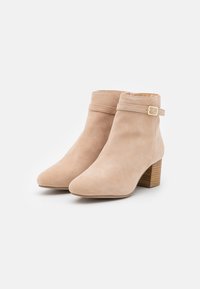 Bottes hauteur cheville en daim beige avec bout rond, talon bloc, et une sangle décorative ornée d'une boucle dorée autour de la cheville.
