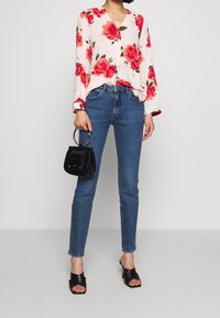 Blouse rose clair avec imprimé floral rouge, manches longues et décolleté en V. Porté avec un jean bleu ajusté et un sac à main noir. Sandales noires à glissière.