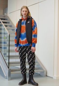 Crna podstavljena prsluk preko narančaste i plave majice s uzorkom vezivanja, uparen s crnim hlačama koje imaju bijeli print "JORDAN", i crnim čizmama do gležnja.