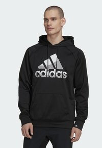 Felpa nera con cappuccio caratterizzata da un grande logo Adidas bianco con un design screziato. Ha una tasca a marsupio e un cappuccio regolabile con polsini a coste.