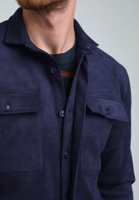 Camicia in suede blu navy con colletto, chiusura a bottoni e due tasche sul petto con pattina. Texture morbida e finitura opaca.