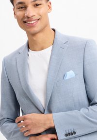 Lichtblauwe blazer van textuurstof, met een enkele borstzak en een lichtblauw pochet, en vierknopenmanchetten. Gecombineerd met een wit T-shirt.