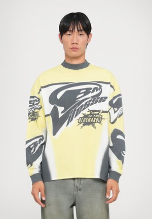 Yellow sweatshirt with graphic black and white designs, ribbed collar, and long sleeves. Paired with light gray, slightly distressed jeans.