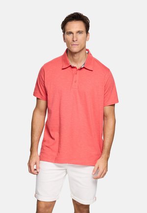 Hombre con polo de manga corta color coral y pantalones cortos blancos con dobladillo enrollado, de pie contra un fondo liso.