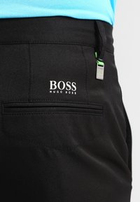 Svarta byxor med en slät yta, med en bakficka och en vit "BOSS HUGO BOSS"-logotyp. Inkluderar en grön detalj på dragkedjan.