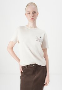 Kremfarget bomull t-skjorte med korte ermer og et brystmotiv som har et design, kombinert med et brunt denimskjørt med en frontlomme.