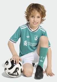 Teal Liverpool FC majica s črnimi in belimi črtami, bele hlače in zelene nogavice. Črni copati; otrok drži črno-belo nogometno žogo.