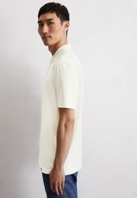 Lichtgekleurde rompershirt met korte mouwen van een textuurstof, met een kraag en zijsplitten, gedragen door een man die iets naar de zijkant is gekeerd.