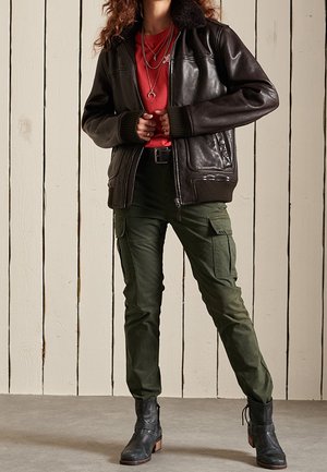 Ruskea nahkainen bomber-takki mustalla turkiskauluksella, alla punainen paita, yhdistetty oliivinvihreisiin cargo-housuihin ja mustiin nilkkureihin.