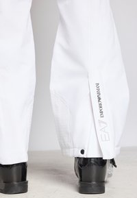 Λευκό αδιάβροχο παντελόνι με υφασμάτινες πλαϊνές πιέτες, φερμουάρ και λογότυπο στο πόδι. Μερικώς ορατές μαύρες γυαλιστερές μπότες.