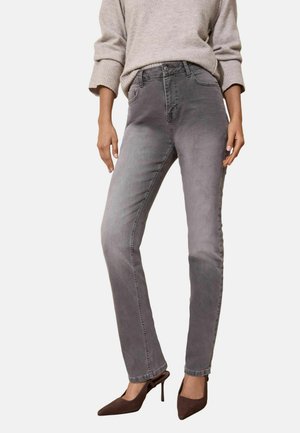 Woman wearing faded gray slim-fit jeans, beige sweater with rolled sleeves, and brown pointed high-heeled shoes standing.