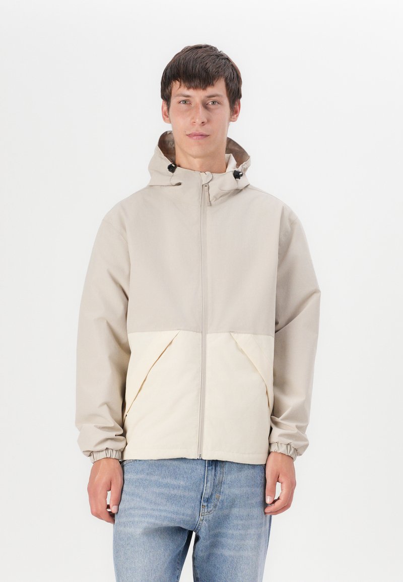 Lichte, beige jas met een crèmekleurig onderpaneel; heeft een capuchon, een rits aan de voorkant, elastische manchetten en zijzakken. Gladde, waterafstotende stof.