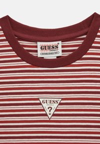 Czerwono-biała koszulka w paski z ryżowanym okrągłym dekoltem. Z przodu znajduje się trójkątny naszywka "Guess Originals".