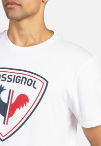 Camiseta de algodón blanca con cuello redondo, presenta un logotipo rojo y negro dentro de un diseño de escudo en el frente. Mangas cortas.