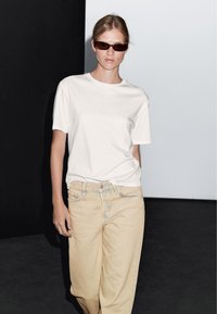 Biała koszulka z okrągłym dekoltem zestawiona z jasnobeżowymi, luźnymi dżinsami; model noszący ciemne, wąskie okulary przeciwsłoneczne w minimalistycznym otoczeniu.