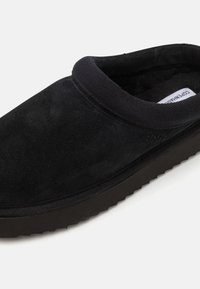 Zapato negro de ante tipo slip-on con punta redondeada, cuello elástico y suela de goma gruesa. Textura suave con acabado liso y minimalismo en la marca.