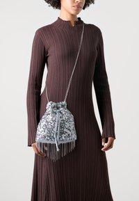 Hopeinen paljettinen nyörilaukku, jossa on hapsureunus ja ketjuhihna. Laukulla on pehmeä, teksturoitu pinta ja pyöreä muoto.