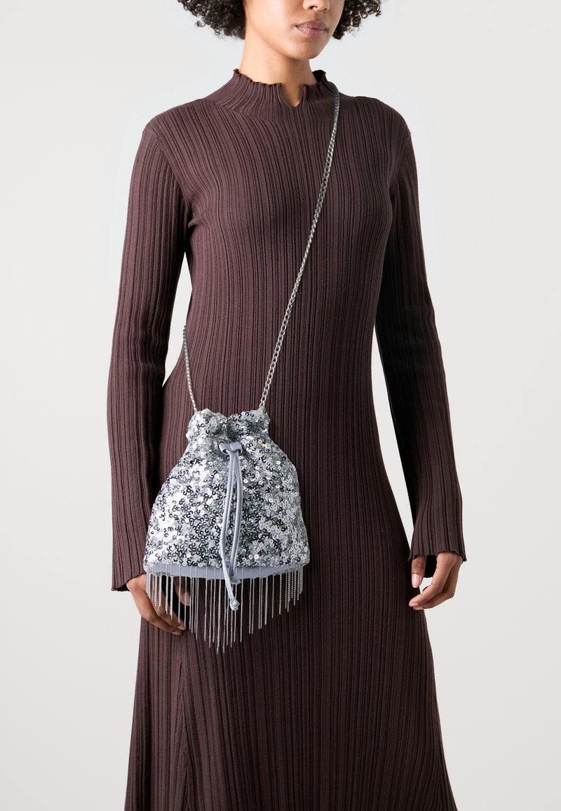 Hopeinen paljettinen nyörilaukku, jossa on hapsureunus ja ketjuhihna. Laukulla on pehmeä, teksturoitu pinta ja pyöreä muoto.