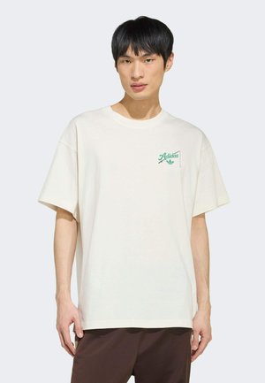 Jeune homme portant un t-shirt blanc ample Adidas avec un logo vert sur la poitrine et un pantalon marron foncé, debout devant un fond uni.