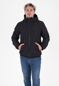 Man in een zwarte hoodie met zijn handen in de zakken en blauwe jeans, recht vooruit kijkend tegen een witte achtergrond.