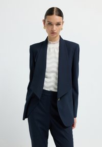 Žena s slicked-back vlasy, oblečená ve zlatých náu�šnicích, námořnickém pruhovaném obleku a bílé vzorované halence na čistém bílém pozadí.