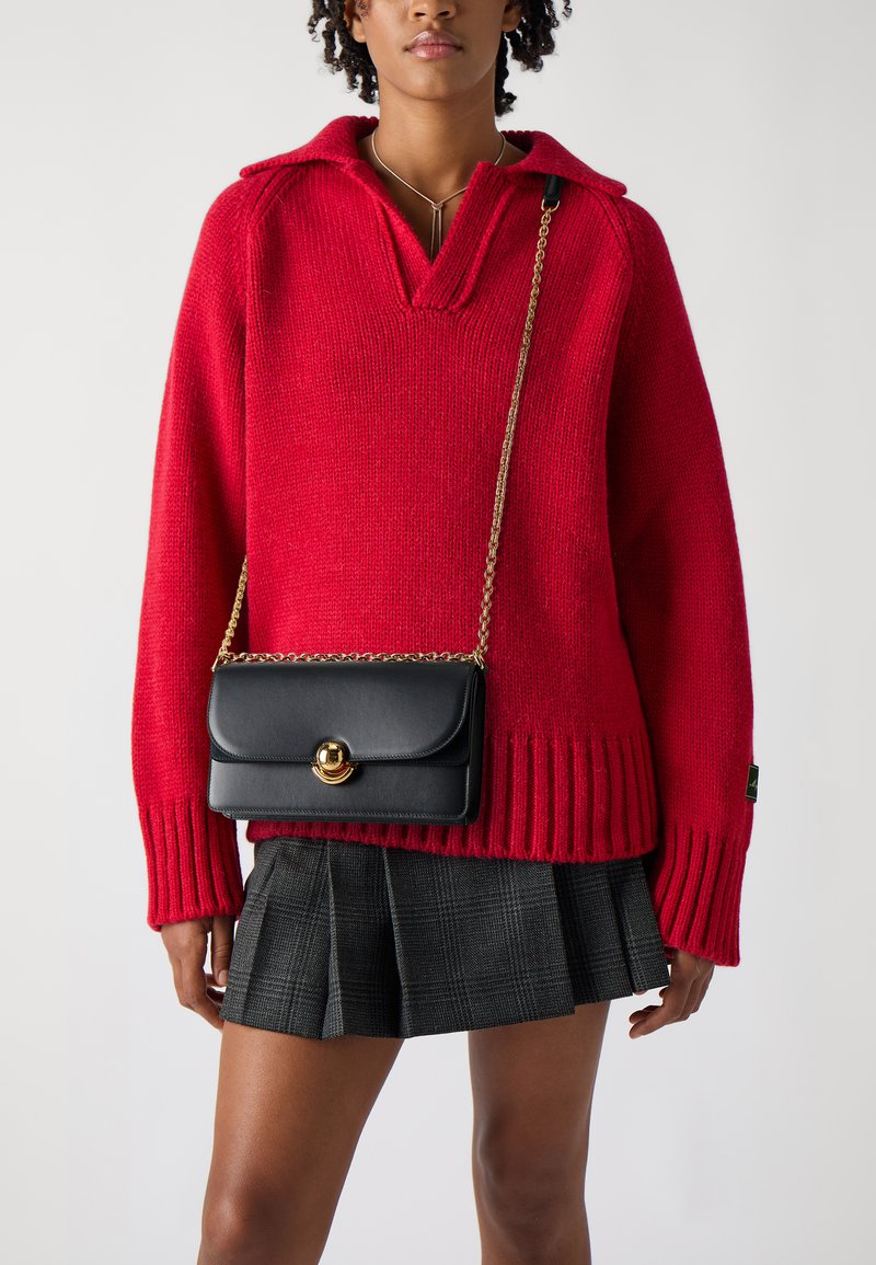 Maglione rosso lavorato a maglia con colletto, abbinato a una borsa a tracolla in pelle nera con chiusura in metallo dorato e una gonna a quadri.