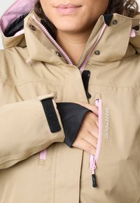 Beige jakke med lyserød lynlås og sort velcro på ærmet, hånd der rækker ind i det forreste brystlomme med teksten "Swedemount" synlig.