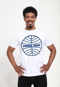 Bílá bavlněná tričko s velkým modrým kruhovým logem, na kterém je v tučném písmu uprostřed napsáno "PAN AM". Krátké rukávy.