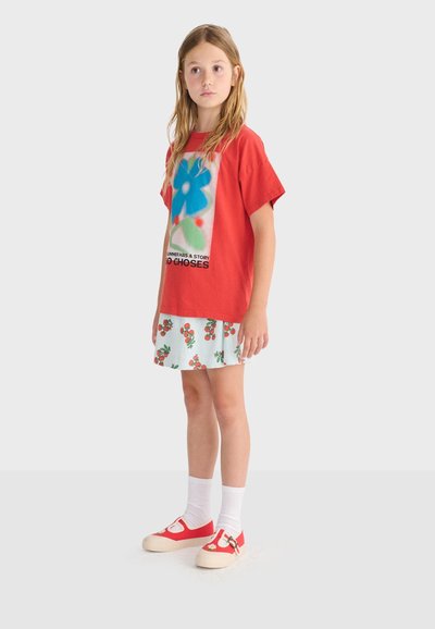 Jeune fille portant un t-shirt graphique rouge, une jupe à imprimé cerises, des chaussettes blanches et des chaussures rouges avec des détails floraux, debout devant un fond uni.