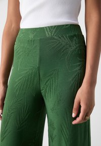 Pantalon vert à jambes larges en tissu lisse, avec un motif subtil de feuilles embossé. La taille est élastique, offrant un ajustement confortable.