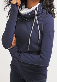 Kvinna som bär en marinblå tröja med mönstrad cowl-hals och långa ärmar, med armarna korsade och en hand nära ansiktet.