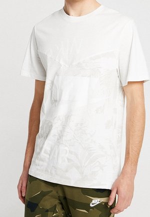 Homme portant une chemise blanche à manches courtes avec des motifs floraux subtils et un short camouflage vert Nike sur un fond uni.
