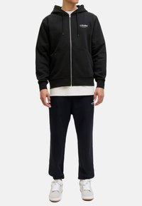 Sweat à capuche noir avec fermeture éclair, cordons de serrage et poches avant, porté sur une chemise blanche, associé à un pantalon de survêtement noir et des baskets blanches.