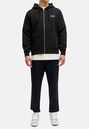 Zwarte zip-up hoodie met trekkoorden en voorzakken, gedragen over een wit shirt, gecombineerd met zwarte sweatpants en witte sneakers.