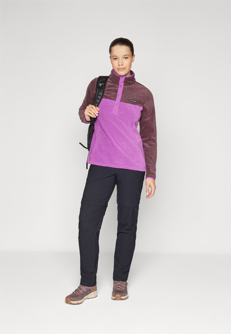 Fleece pullover in paars en bruin met een rits-kraag en knopen aan de voorkant, gecombineerd met zwarte cargo-broeken en bruine wandelschoenen.